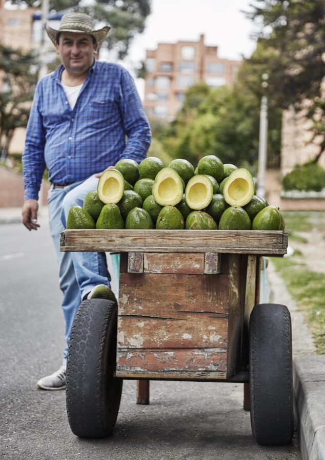 A local Bogota man in a blue shirt and hat stands beside a wooden cart filled with avocados on a vibrant street, reminiscent of Central America's bustling markets.