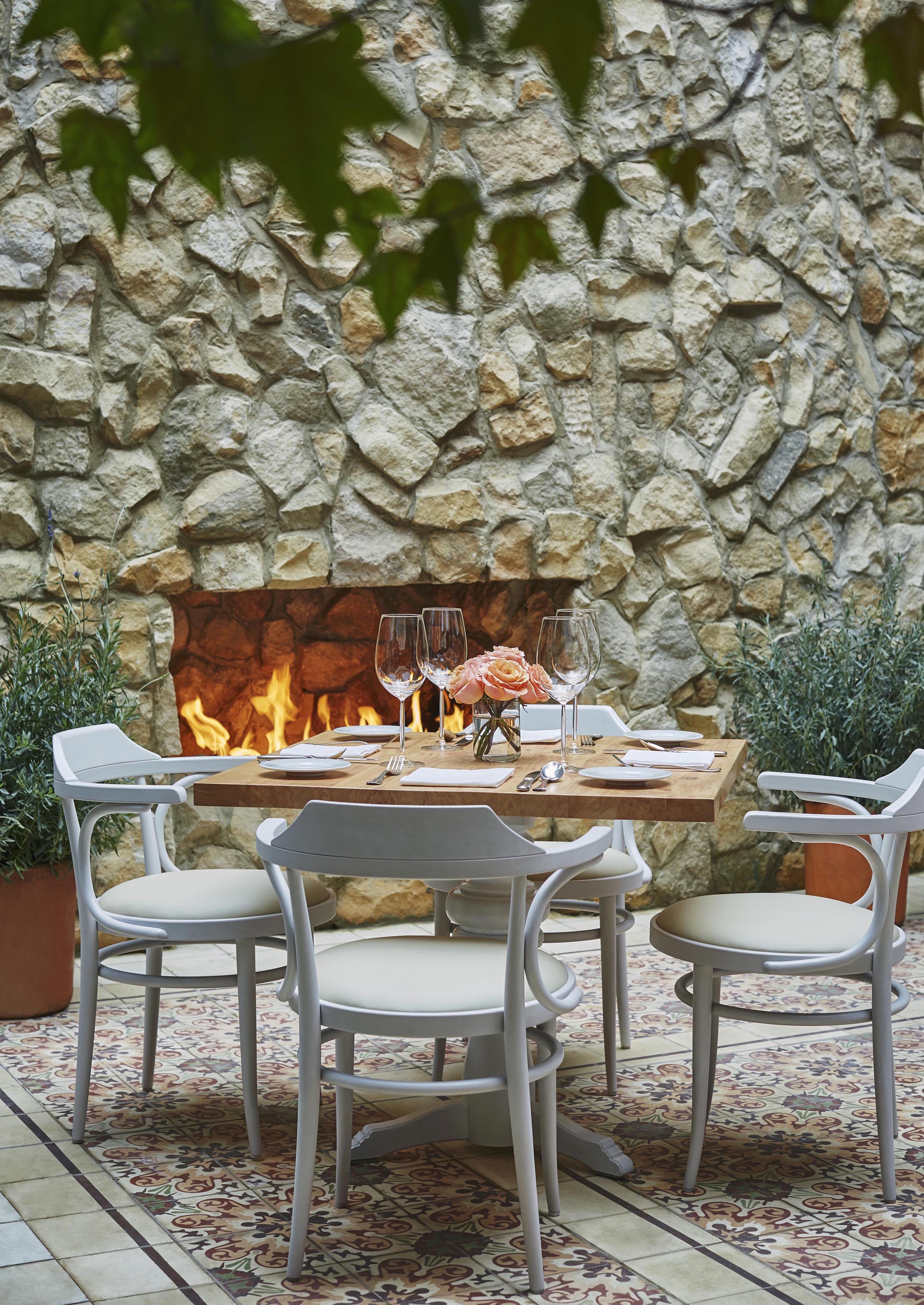 Outdoor dining setup within destination luxury hotel, Four Seasons Bogota, with four white chairs around a wooden table, set with glassware and a floral centerpiece. A stone wall with a lit fireplace evokes the cozy charm of Central America in the background.