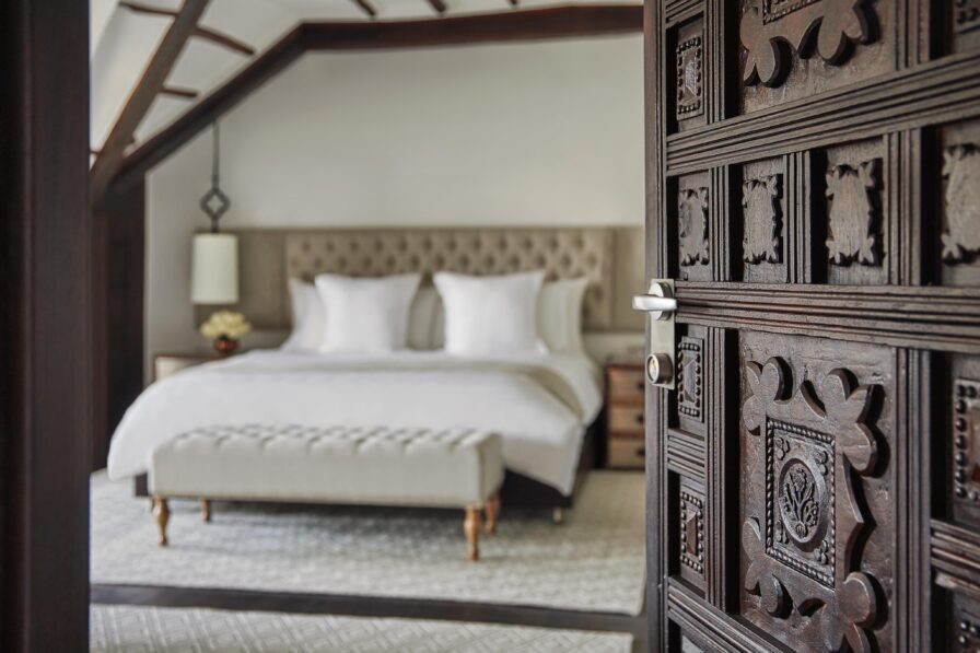 An ornate wooden door opens to a elite luxury hotel suite bedroom of Four Seasons Bogota with a large bed, tufted headboard, and white bedding. A bench sits at the foot, while a lamp and nightstand add charm. Rich textiles evoke the vibrant spirit of Central America, with a colorful rug covering the floor.