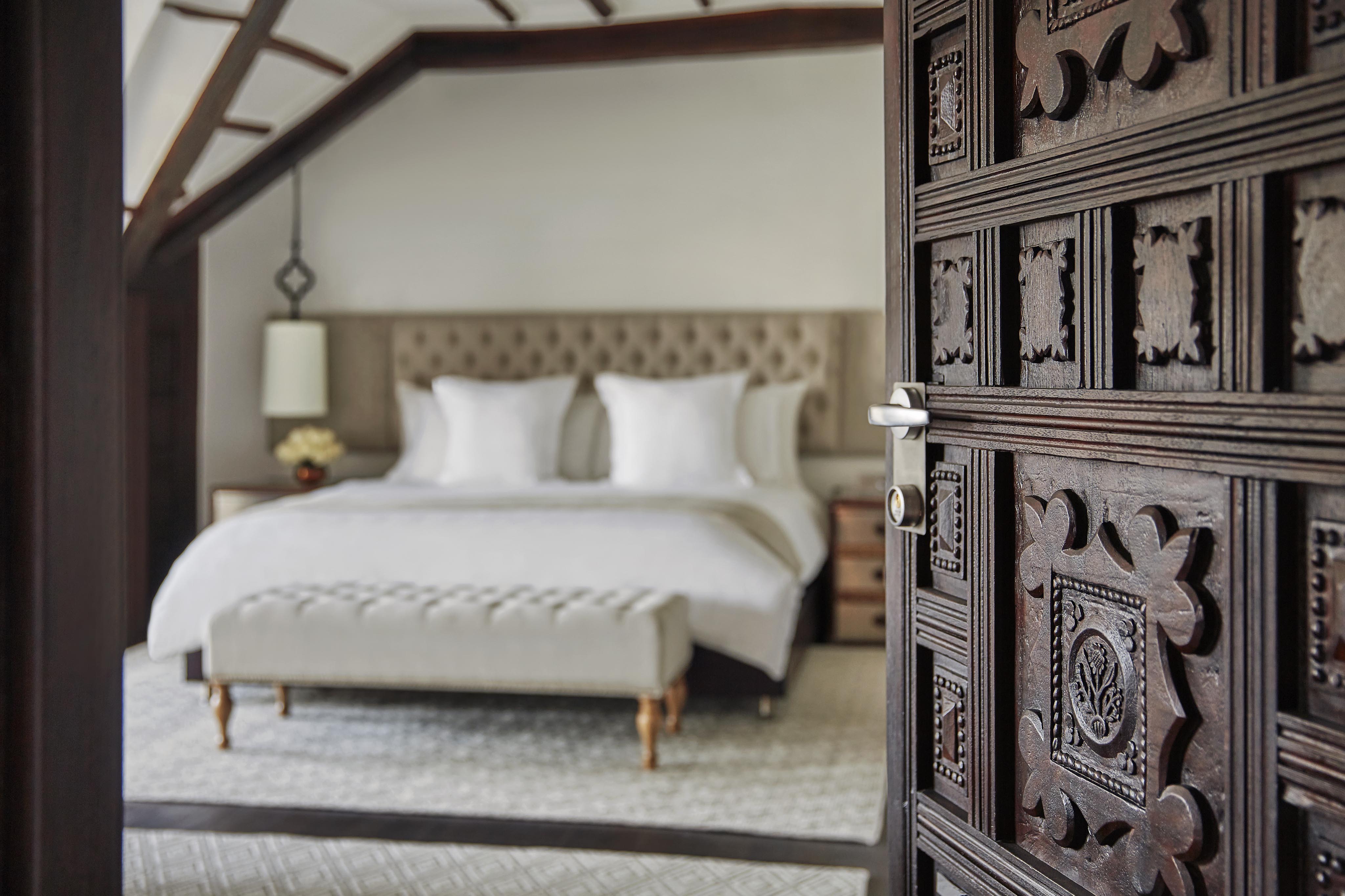 An ornate wooden door opens to a elite luxury hotel suite bedroom of Four Seasons Bogota with a large bed, tufted headboard, and white bedding. A bench sits at the foot, while a lamp and nightstand add charm. Rich textiles evoke the vibrant spirit of Central America, with a colorful rug covering the floor.
