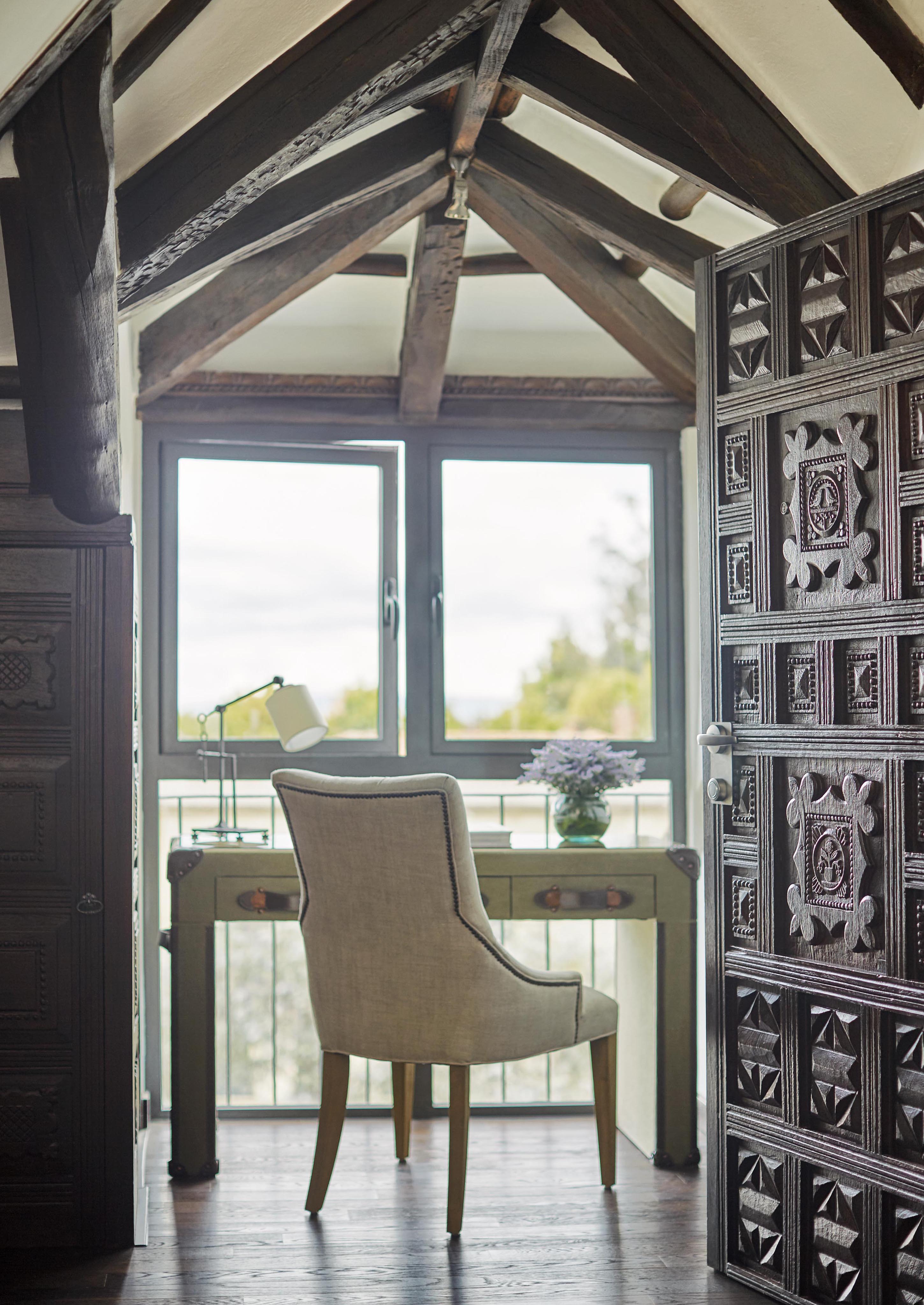 Cozy workspace within the Four Seasons Bogota destination luxury hotel suite featuring wooden beams, large window, desk, chair, lamp, and potted plant. Sunlight illuminates the area, highlighting ornate wooden door details reminiscent of South American craftsmanship.