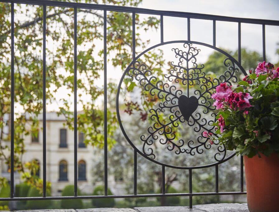 Wrought iron balcony railing with heart and floral design, pink flowers in a terracotta pot on the right, echoing vibrant scenes of South America, with blurred views of a building and trees in the background.