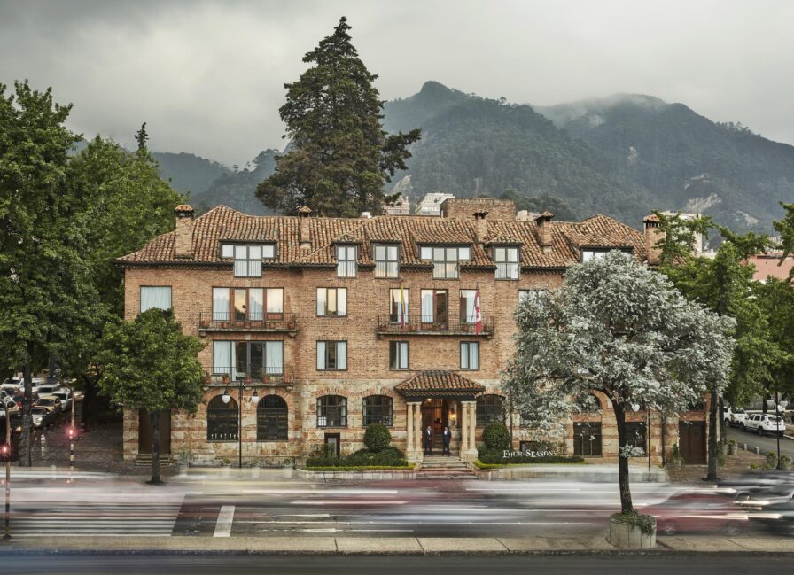 Historic brick exterior architecture of luxury hotel Four Seasons Casa Medina with a tiled roof, set in a lush green area with trees. Mountains and clouds are visible in the background. Cars pass by on the road in front.
