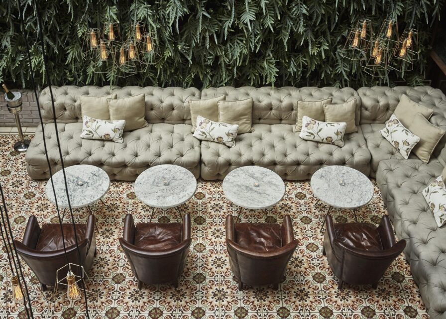 Aerial view of a Four Seasons Casa Medina Bogota lounge area with a long tufted sofa, floral cushions, round marble tables, and leather chairs, set against a wall with lush greenery reminiscent of lush Central American jungles.