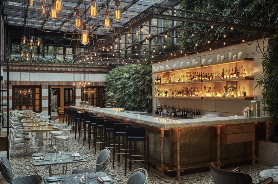 Chic restaurant interior Castanoyles of Four Seasons Casa Medina with a large bar, hanging lights, and lush greenery reminiscent of Central America on walls. Elegant dining tables are set beneath a glass ceiling, offering a modern decor touch that feels both exotic and inviting.