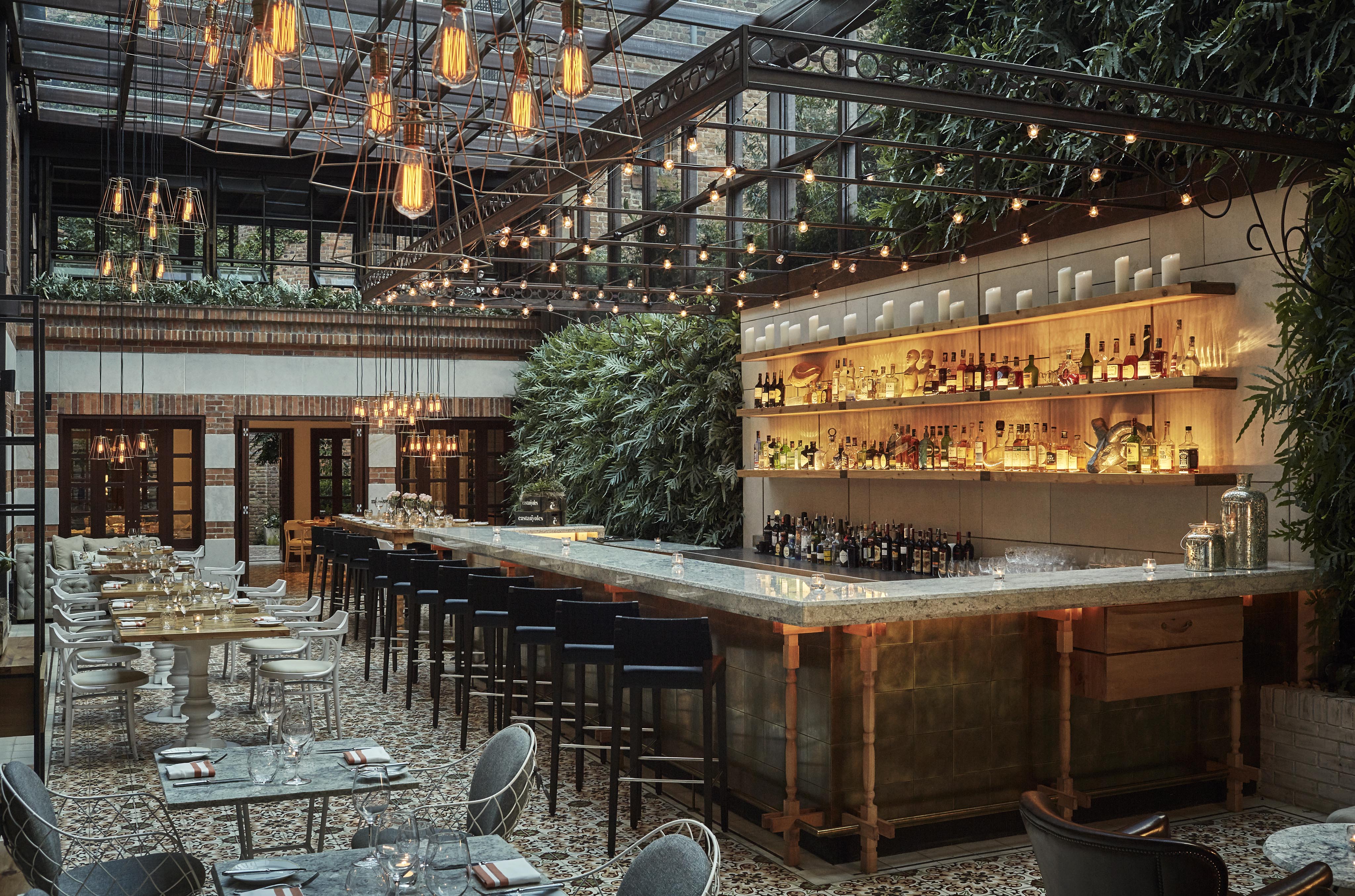 Chic restaurant interior Castanoyles of Four Seasons Casa Medina with a large bar, hanging lights, and lush greenery reminiscent of Central America on walls. Elegant dining tables are set beneath a glass ceiling, offering a modern decor touch that feels both exotic and inviting.