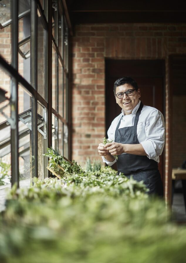 A Four Seasons Casa Medina chef in a white coat and black apron stands by a table with fresh herbs, evoking the rich culinary traditions of destination areas of Central and South America, near large windows in a brick-walled room, smiling as they hold vibrant greens.