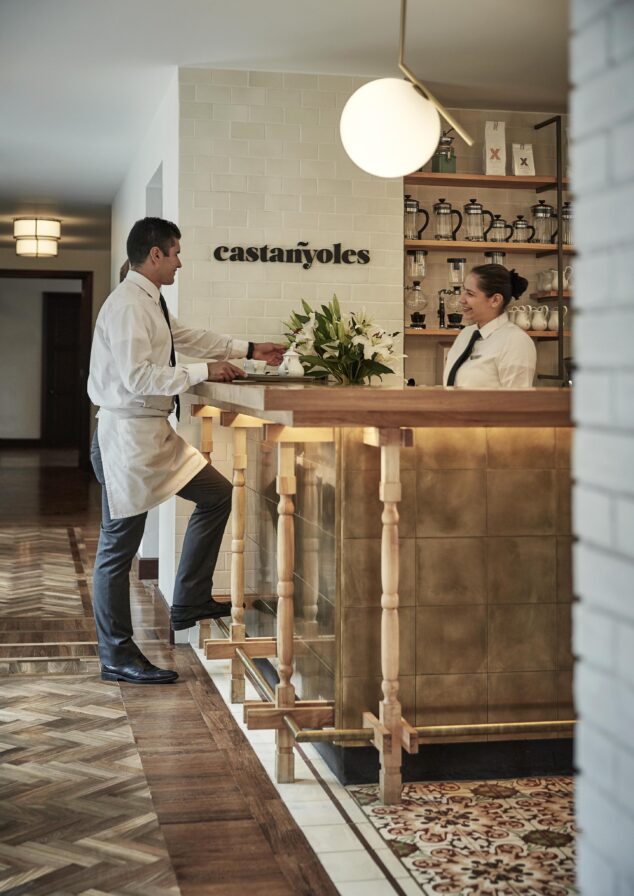A hospitality staff member of Four Seasons Casa Medina destination luxury hotel stands in a white coat and a woman in a white shirt stand at a brass counter in a room with patterned floors, labeled "castañoles," echoing the vibrant cultures of Central and South America.