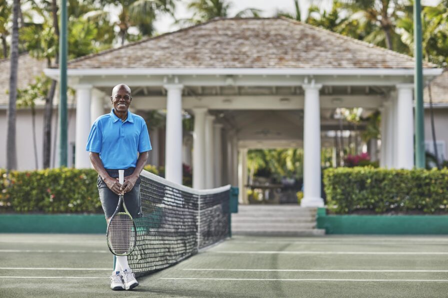 Portrait of the Ocean Club, A Four Seasons resort hospitality staff of in a blue shirt stands on a tennis court holding a racket, with a net in front and a pavilion with columns in the background.