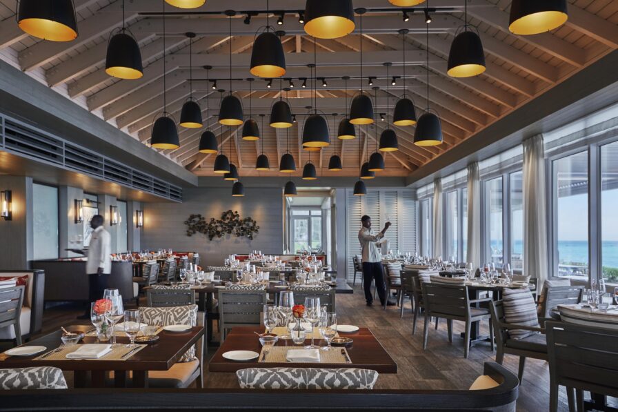 A spacious Ocean Club, A Four Seasons resort restaurant with luxury interior featuring high ceilings, modern lighting, neatly set tables, and large windows revealing a sea view. Staff members are preparing the dining area.