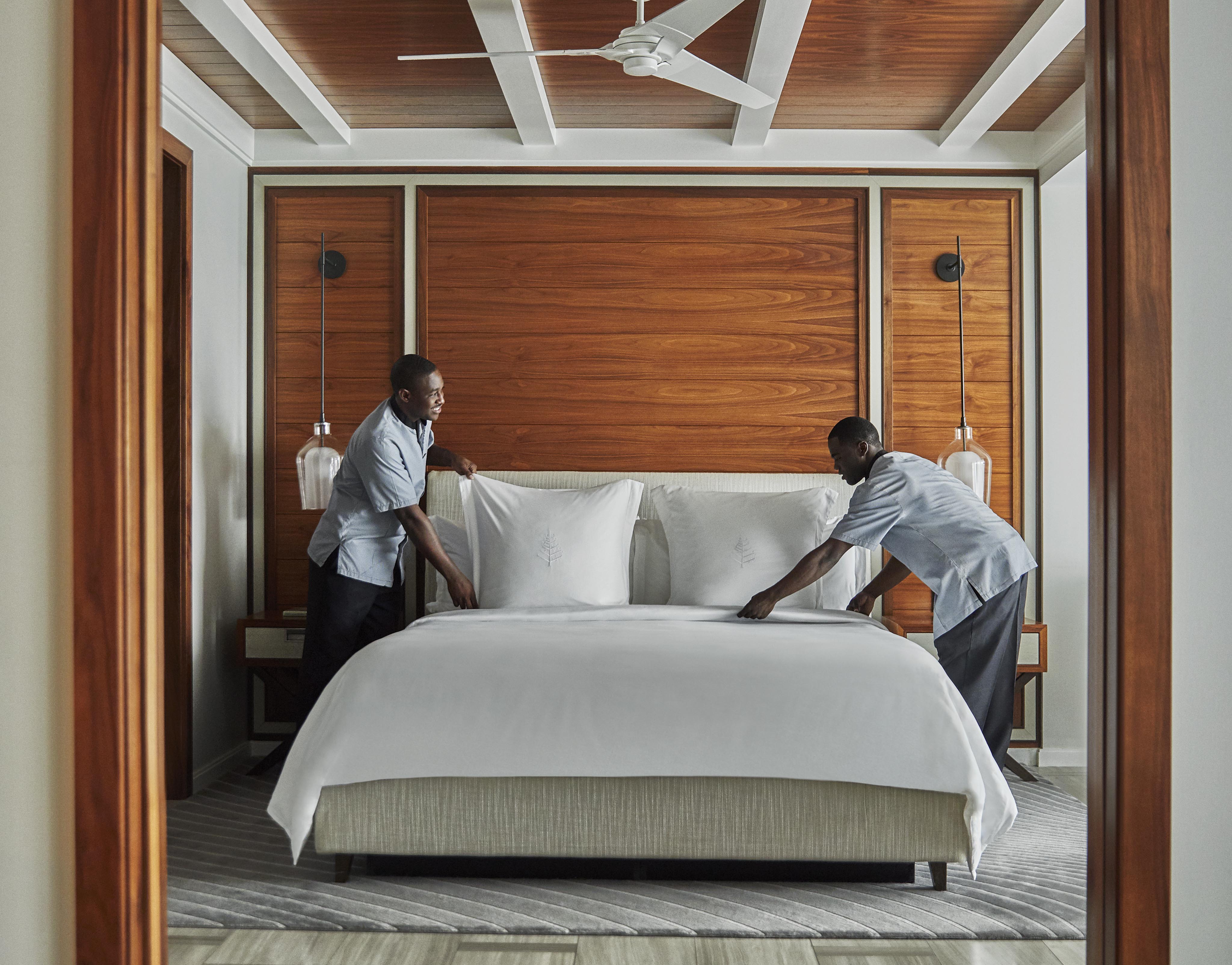 Two Ocean Club, A Four Seasons Resort hospitality staff members in uniforms make a bed with white linens in a room with wooden accents and a ceiling fan.