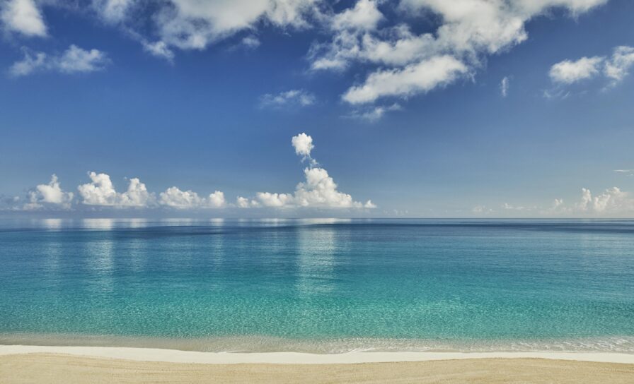 An empty sandy Ocean Club, a Four Seasons resort beach meets a calm, clear turquoise ocean under a bright blue sky with white clouds.