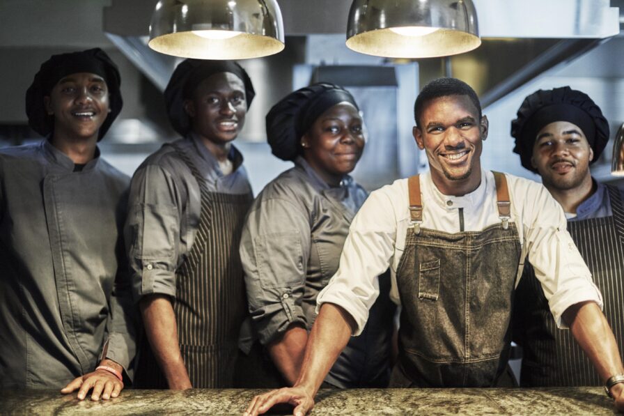 Five chefs of Ocean Club, A Four Seasons resort upscale restaurant "Dune", captured smiling and in uniforms gather in a kitchen, all smiling and standing under hanging lights.