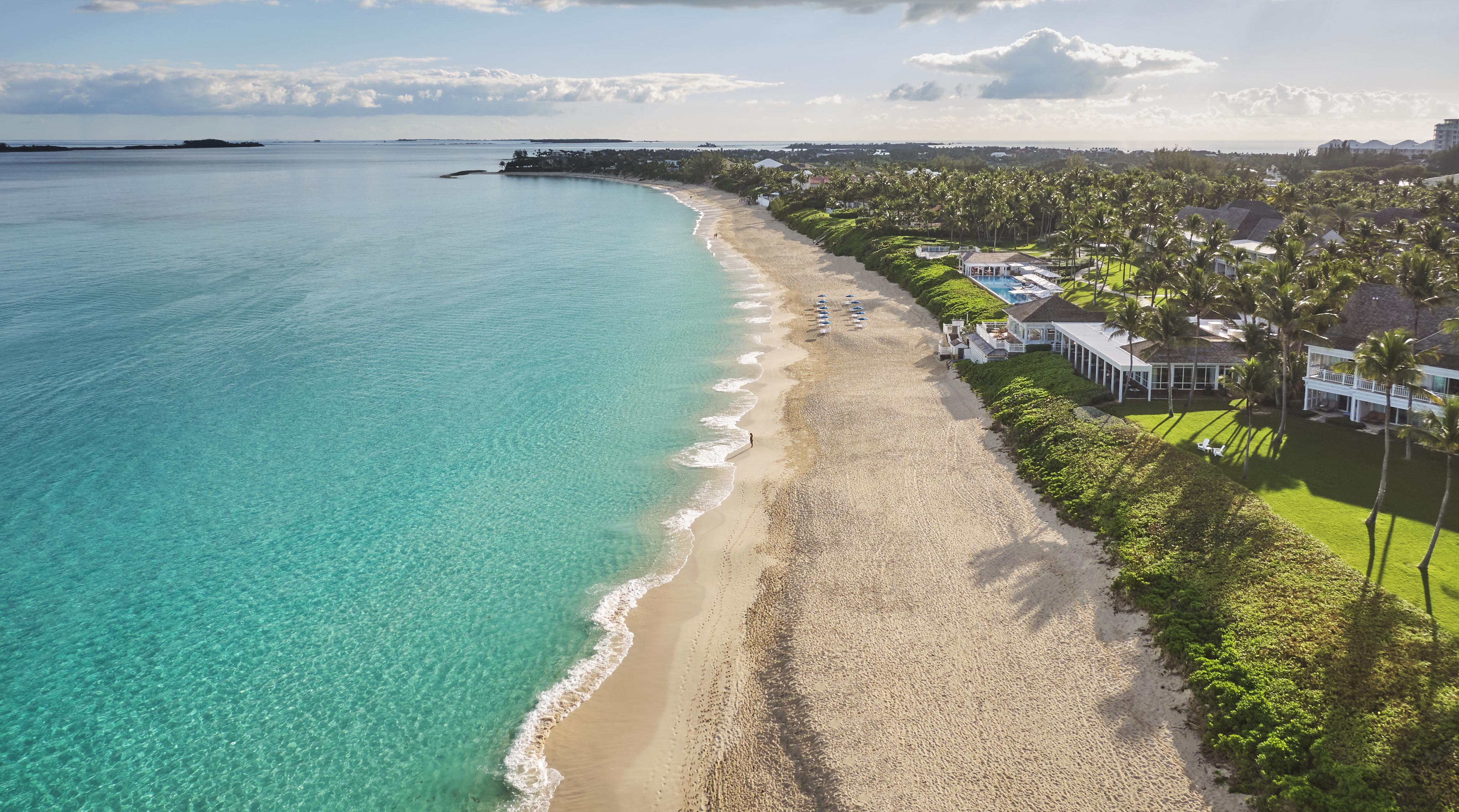 Aerial view of a tropical Bahamas beach, The Ocean Club, A Four Seasons Resort property nestled along the coastline, with turquoise water, gentle waves, and a sandy shore. There are luxury hotel beach chairs, green palm trees, and buildings along the coast under a blue sky with clouds.