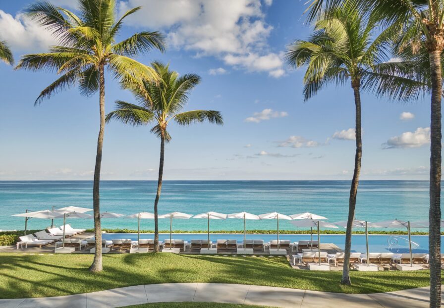 A scenic view of a beachfront Four Seasons Ocean Club resort with palm trees, resort lounge chairs, and luxury hotel umbrellas beside an infinity pool overlooking a turquoise ocean under a blue sky.
