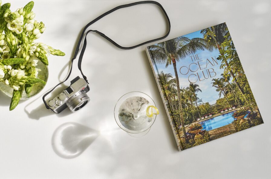 A camera, a magazine titled "The Ocean Club," and a drink with a lemon slice on a white surface. A bouquet of white flowers is on the left. The image is bright with leafy shadows.