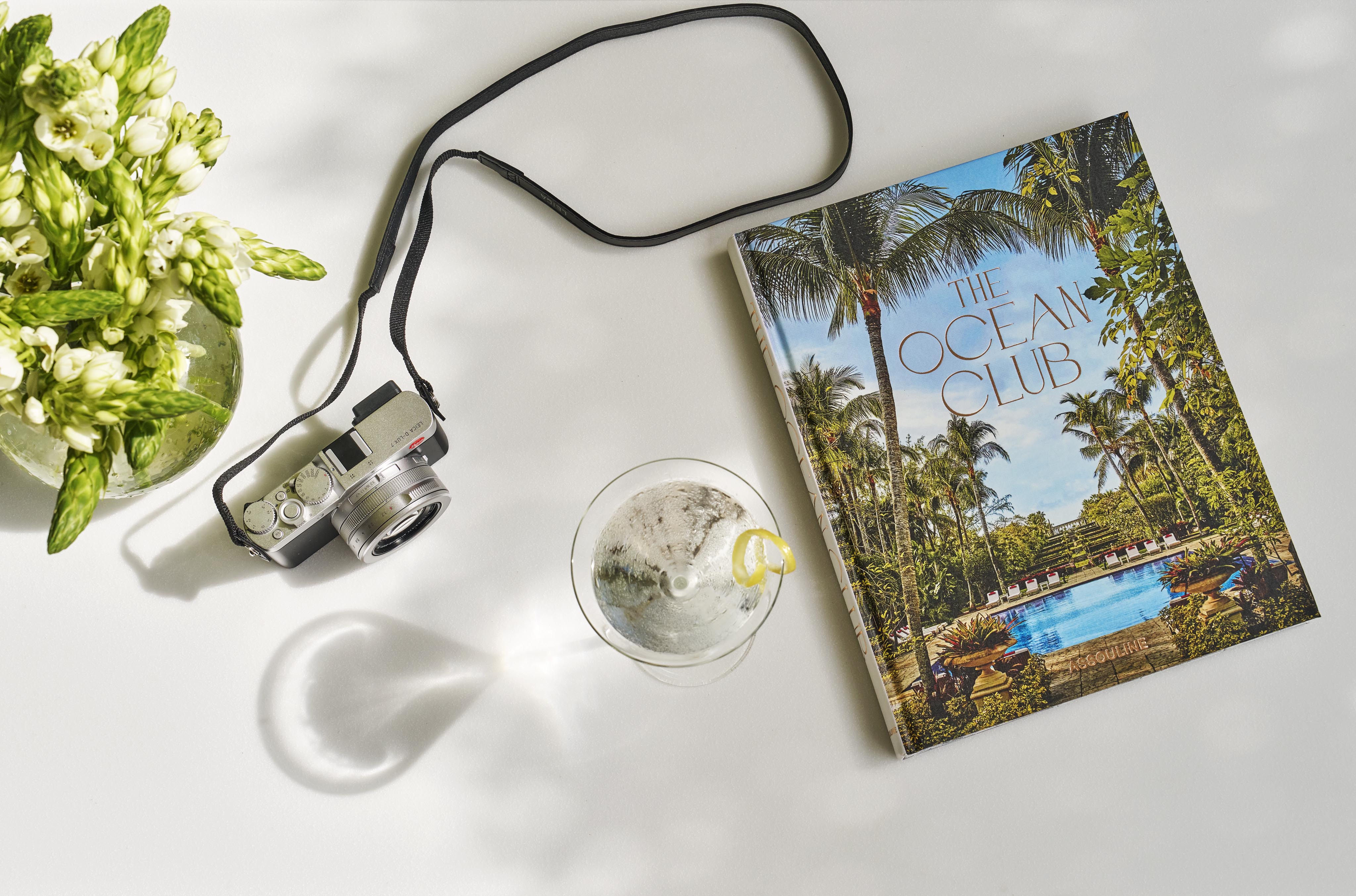A camera, a magazine titled "The Ocean Club," and a drink with a lemon slice on a white surface. A bouquet of white flowers is on the left. The image is bright with leafy shadows.