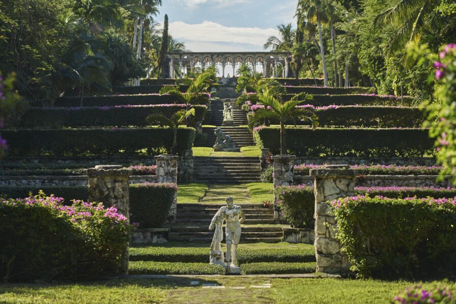 Manicured Paradise Island Versailles garden with a statue and tiered steps leading to an arched structure. Lush greenery and purple flowers surround the scene on a Four Seasons Ocean Club sunny day.