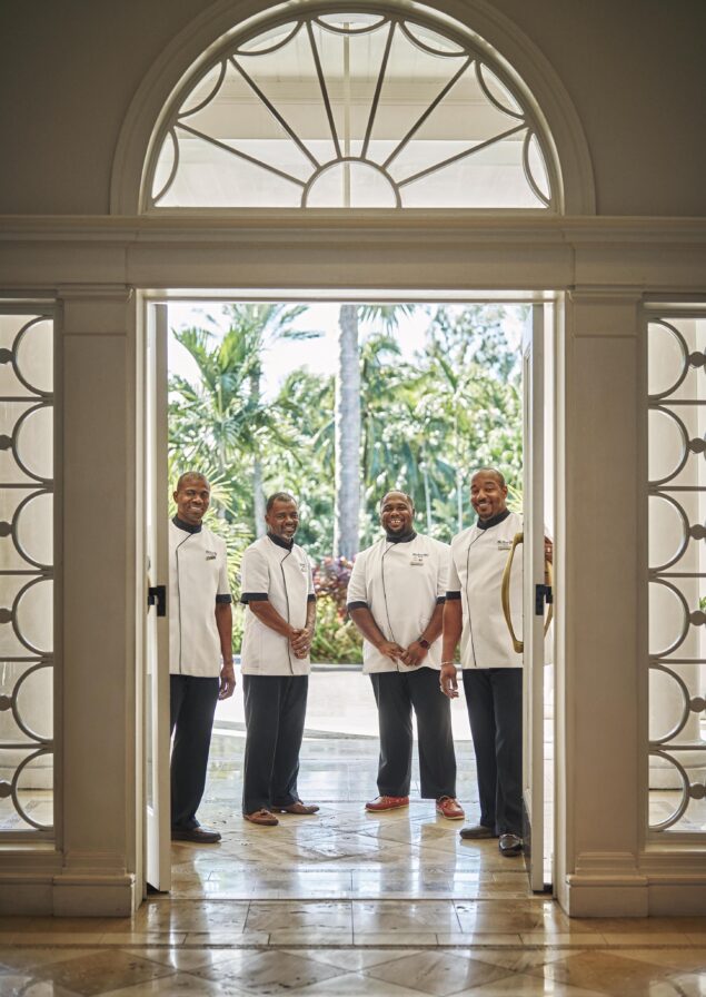 Four hospitality staff in uniform stand smiling in an arched doorway of The Ocean Club, A Four Seasons Resort with glass details, opening to a view of tropical plants and trees.