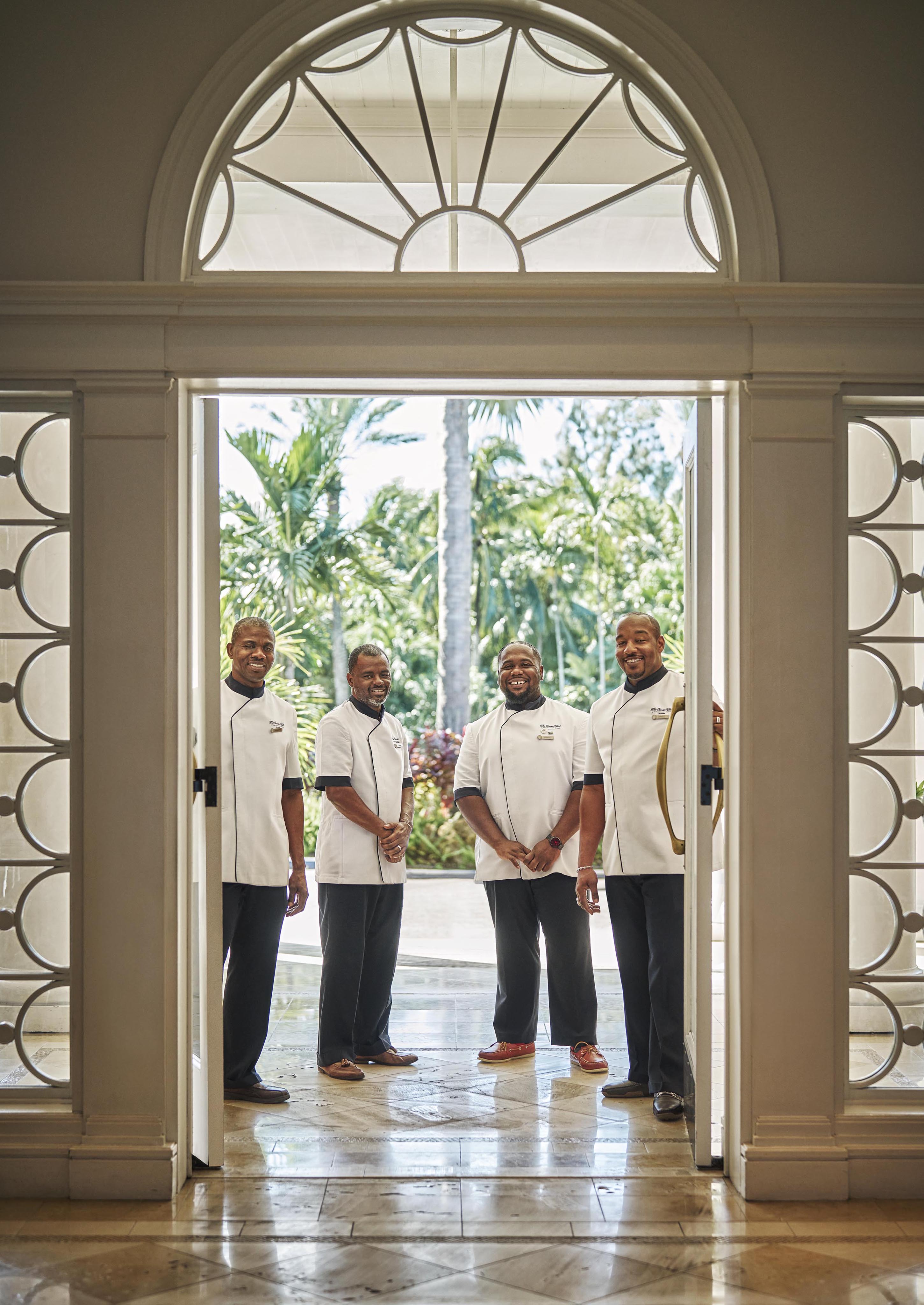 Four hospitality staff in uniform stand smiling in an arched doorway of The Ocean Club, A Four Seasons Resort with glass details, opening to a view of tropical plants and trees.
