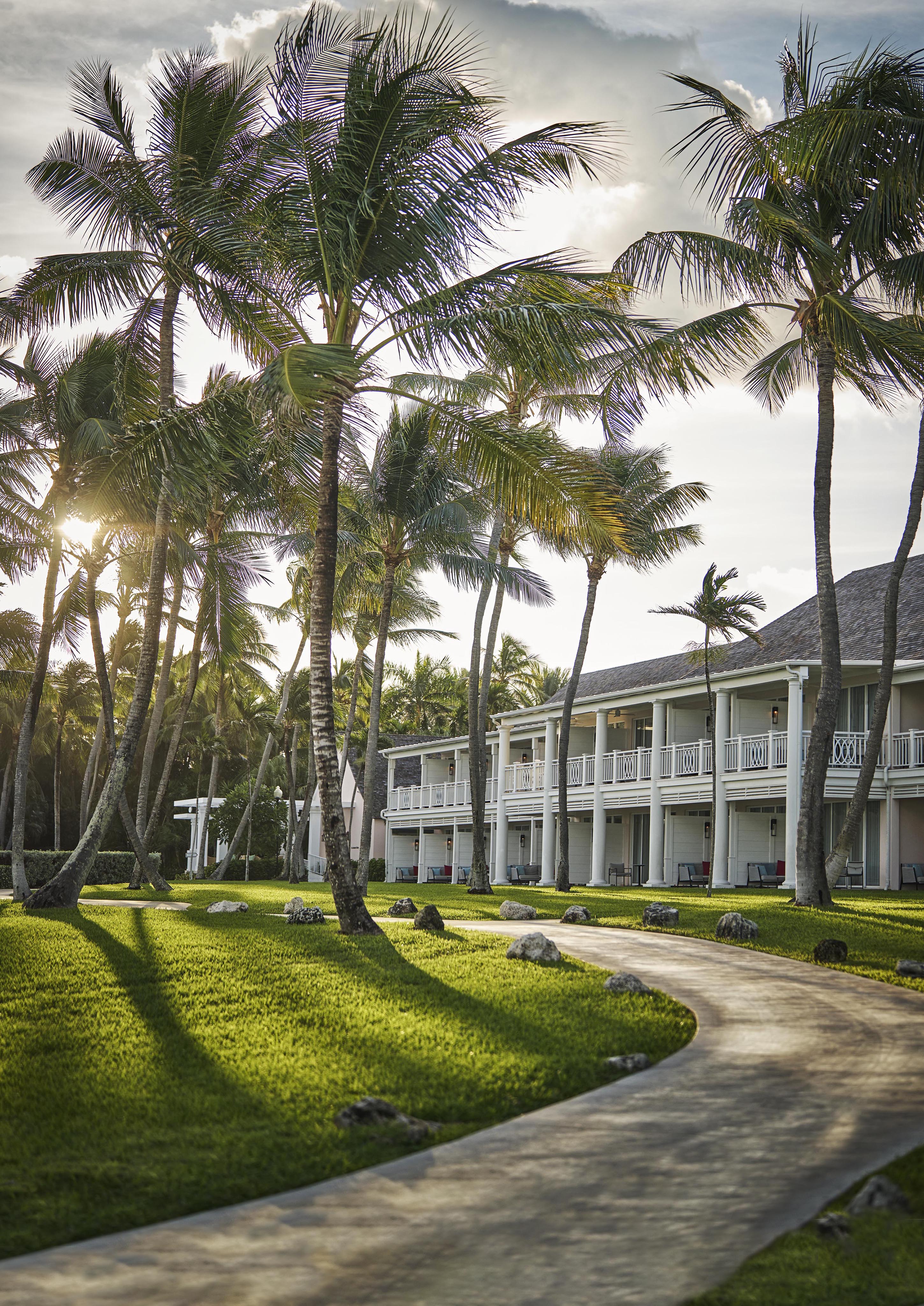 A palm-lined pathway of The Ocean Club, A Four Seasons resort curves through a grassy lawn, leading to a two-story white building under a partly cloudy sky.