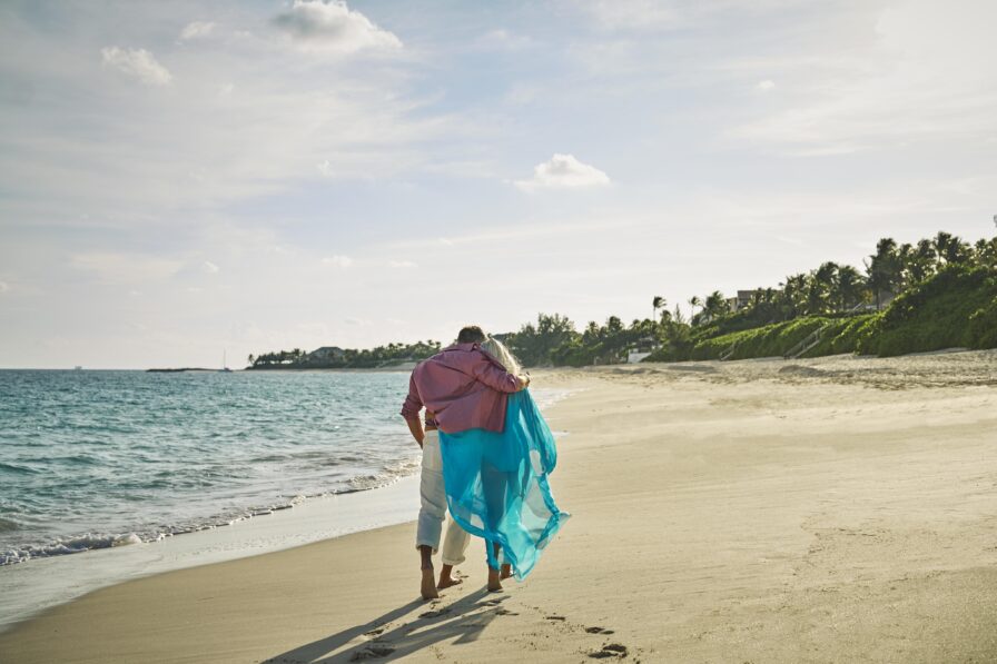 A couple walks barefoot along a sandy beach in front of the Ocean Club, A Four Seasons resort, leaving footprints. The person on the right is draped in a blue scarf, and the sea is calm. Distant houses and greenery line the shoreline.