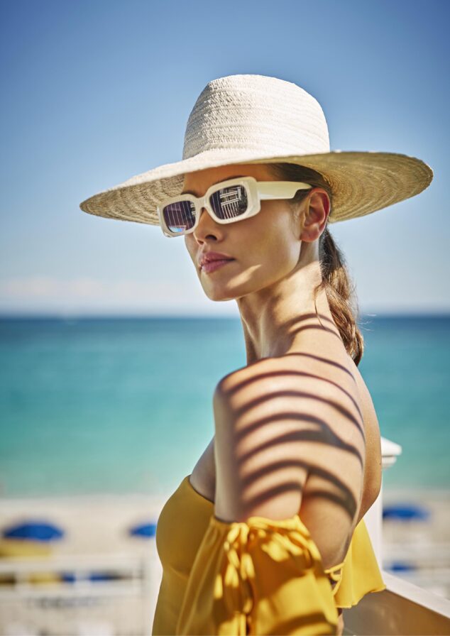 Woman in wide-brimmed hat and sunglasses stands on a beach, wearing a yellow top. The Nassau, Bahamas ocean and blue sky serve as the backdrop.