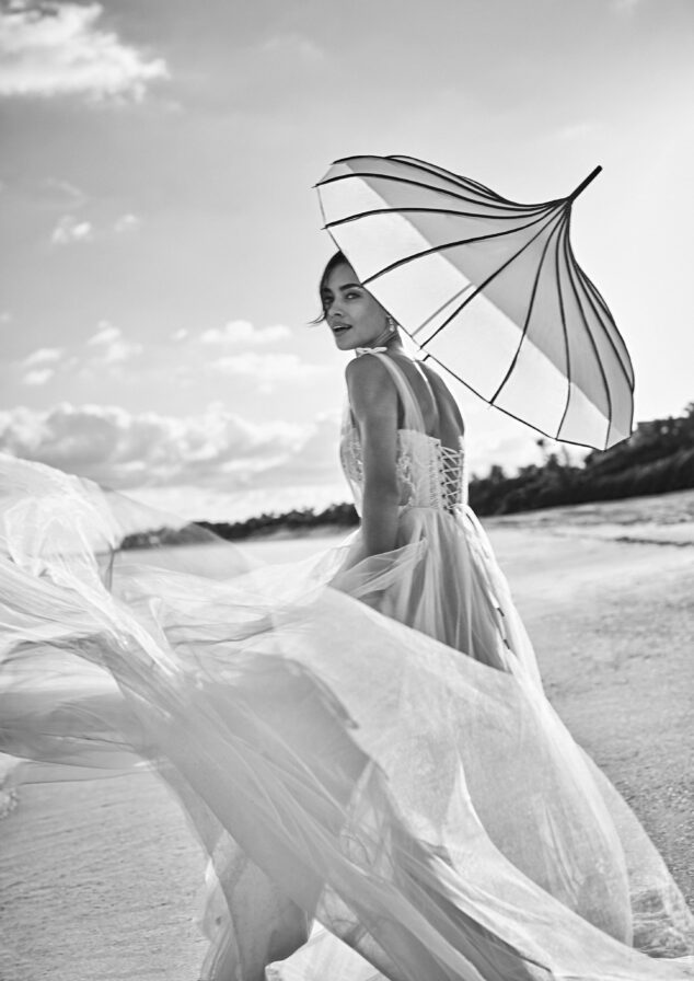 A woman in a flowing dress holds a striped umbrella on a beach. The image is in black and white, with a cloudy Bahamas sky in the background.