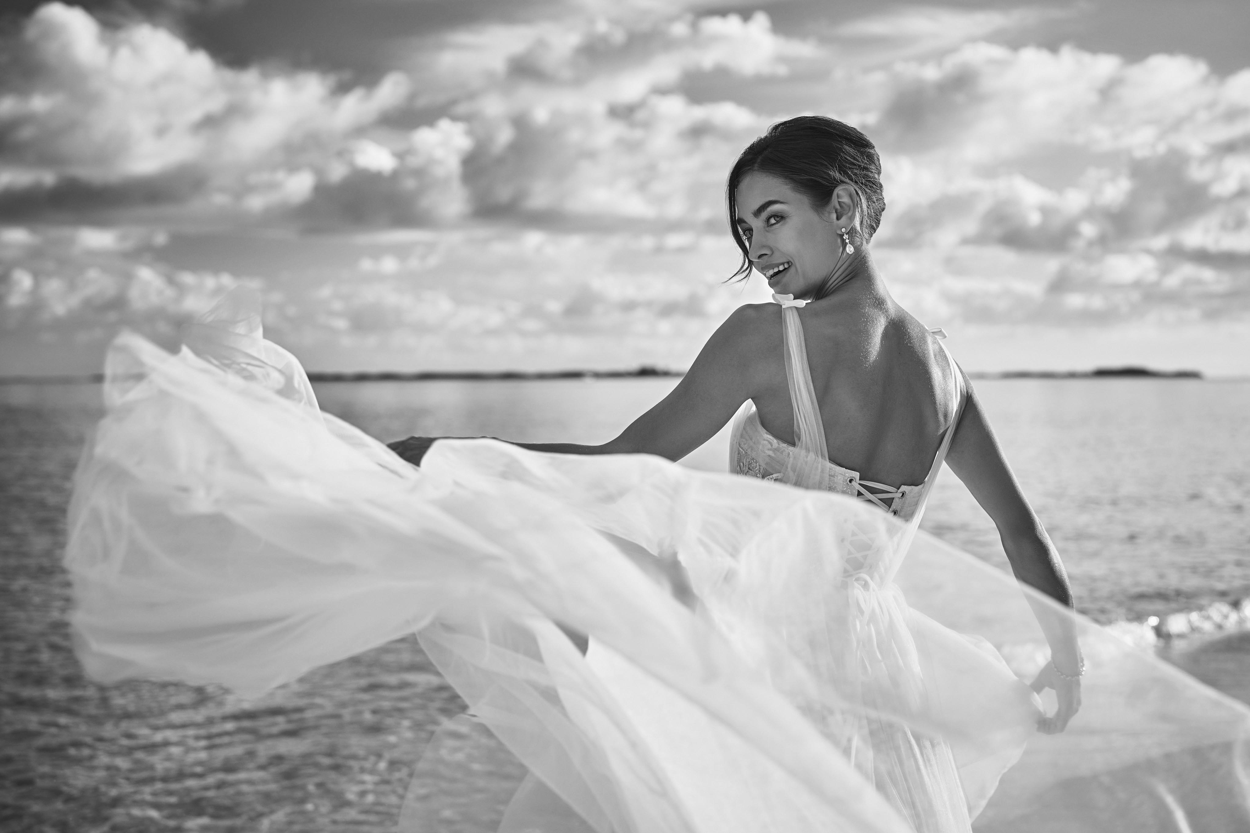 A woman in a flowing dress looks over her shoulder, standing by the water with a cloudy Bahamas sky in the background. Black and white image.
