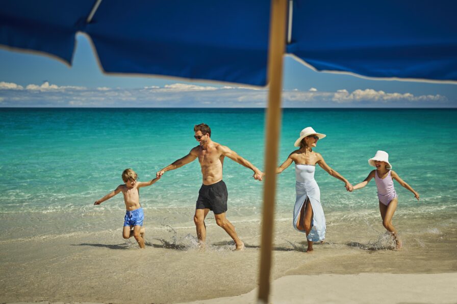 Family of four walks hand in hand along a sandy beach by clear blue ocean water, under a sunny sky with visible blue luxury Ocean Club, A Four Seasons resort hotel umbrella above.
