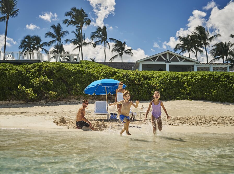 Two children run from the water on a sandy beach in front of The Ocean Club, A Four Seasons resort, with two adults sitting near blue beach umbrellas and a house in the background under a clear sky.