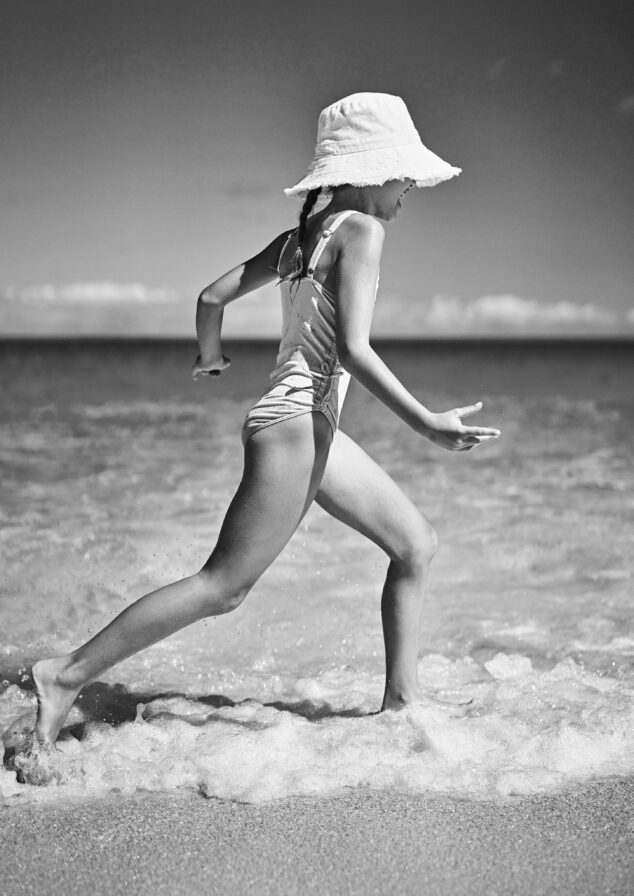 A child wearing a swimsuit and sun hat joyfully runs along the shoreline in front of The Ocean Club, A Four Seasons Resort, splashing water in a black and white beach scene.