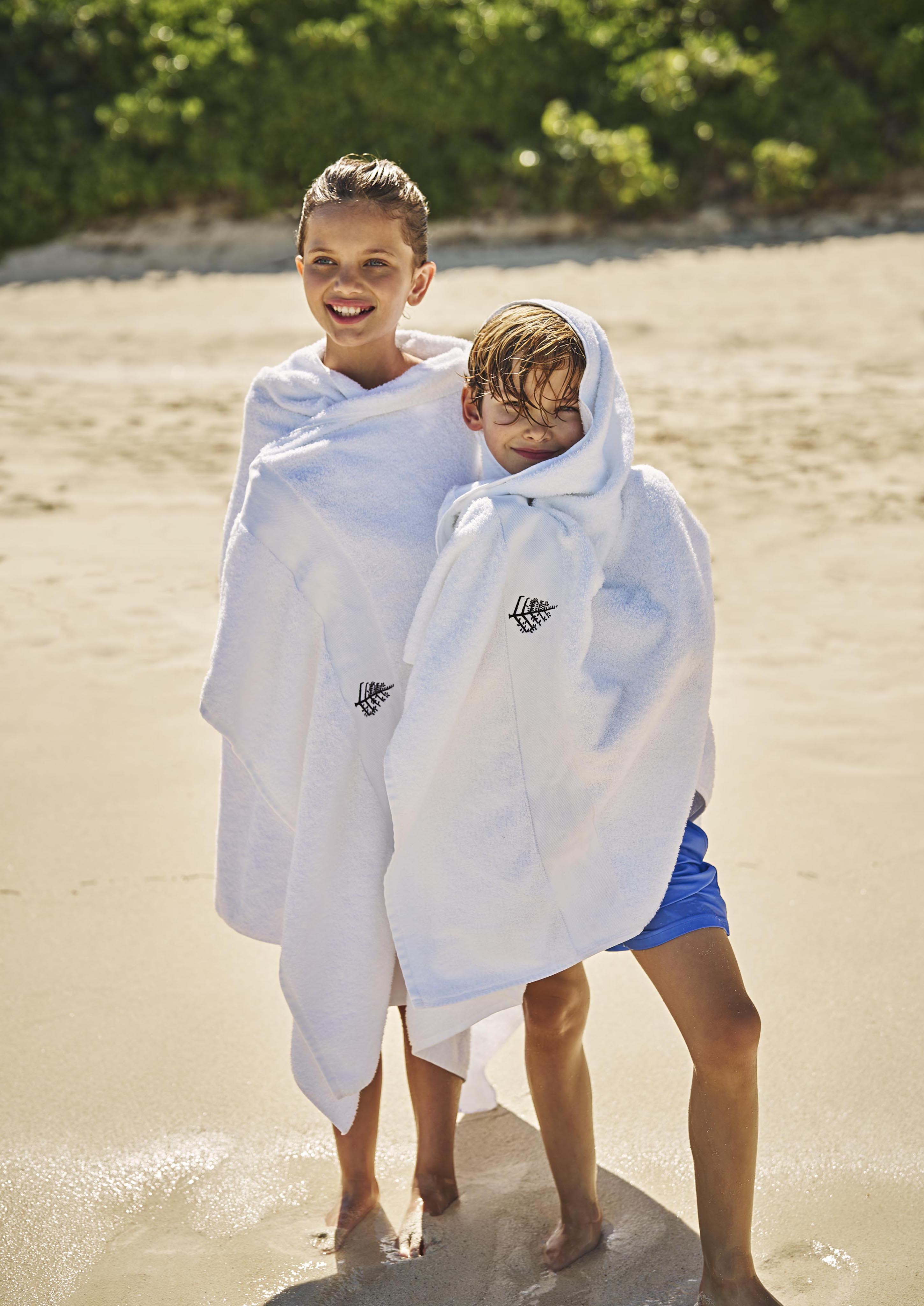 Two children wrapped in white luxury hotel towels stand on a sandy Bahamas beach in front of The Ocean Club, A Four Seasons Resort. One child smiles while the other looks down. Green foliage is visible in the background.