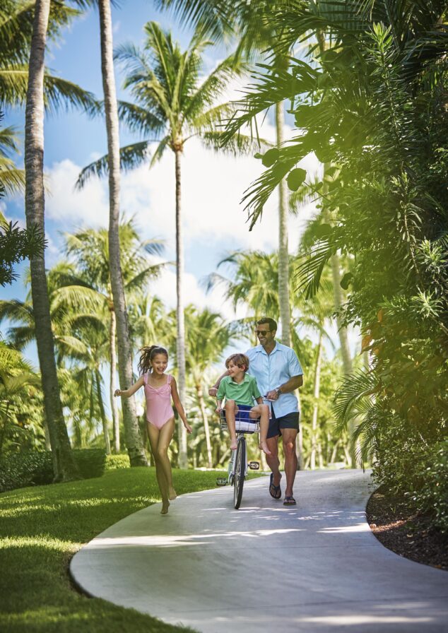 Authentic lifestyle capture of man biking with a child on the handlebars and a girl walking beside them on a Ocean Club, A Four Seasons resort path lined with palm trees.