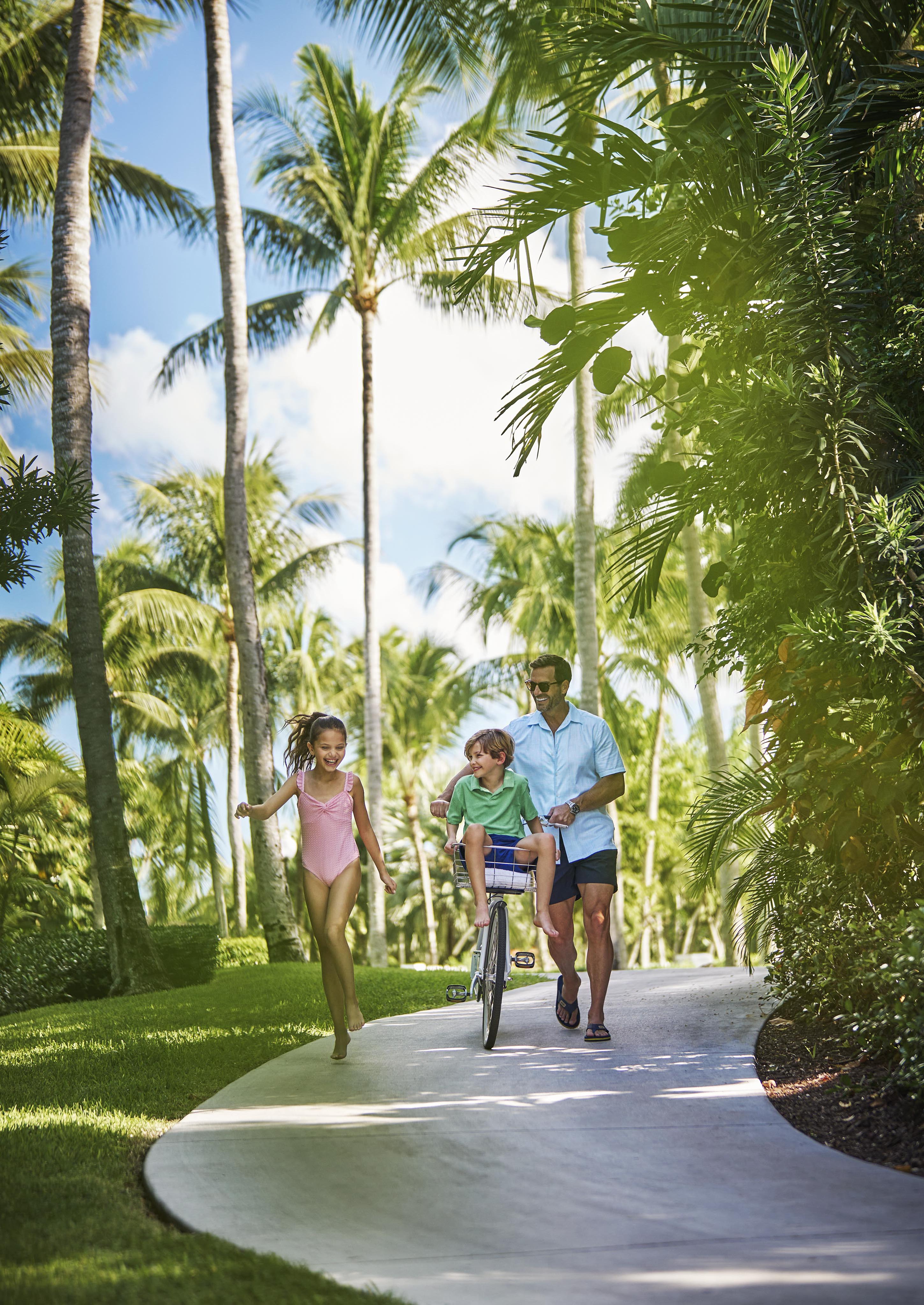 Authentic lifestyle capture of man biking with a child on the handlebars and a girl walking beside them on a Ocean Club, A Four Seasons resort path lined with palm trees.