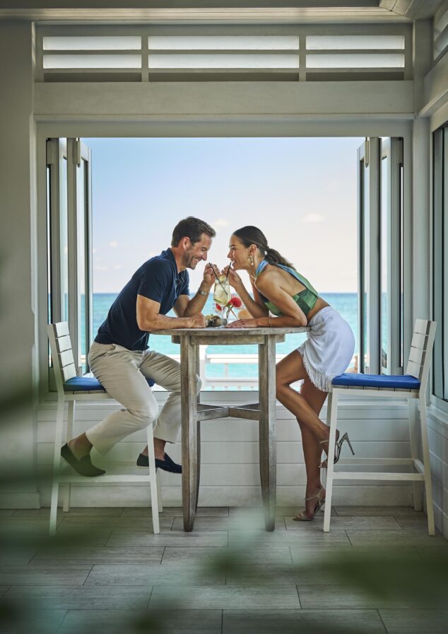 A man and woman smile at each other, sitting at a tall table at The Ocean Club, A Four Seasons Resort, Dune Bar, the couple is next to a large open window with a Paradise Island Bahamas ocean view. They are sharing a drink with a straw.