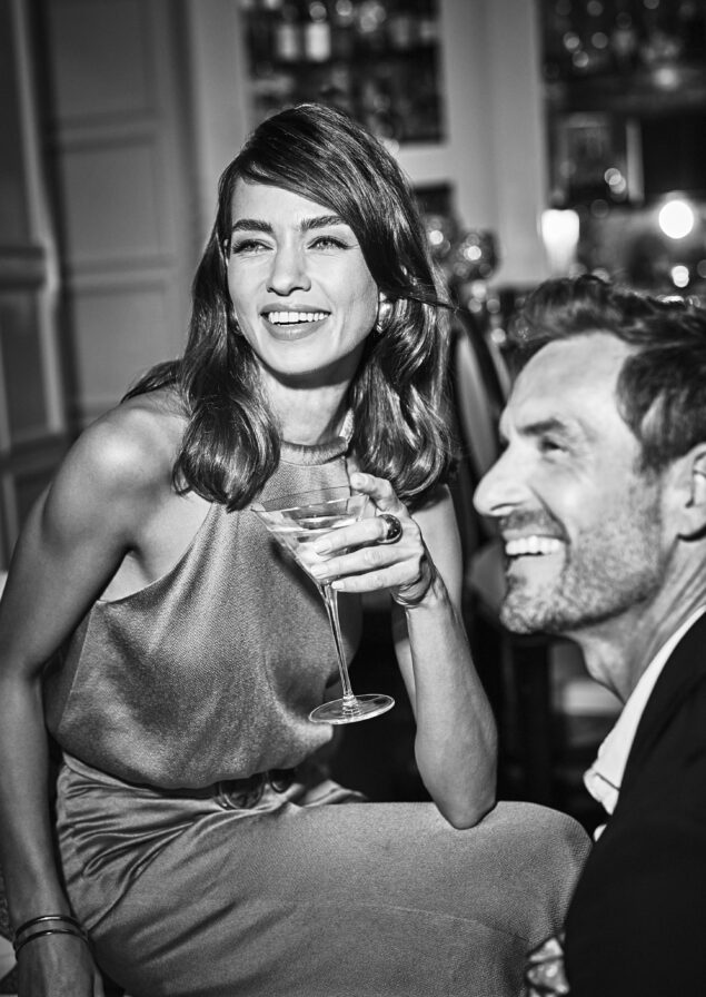 Woman holds a cocktail, smiling. A man in a suit sits nearby, also smiling. They are indoors at a social gathering at the Ocean Club, A Four Seasons resort.