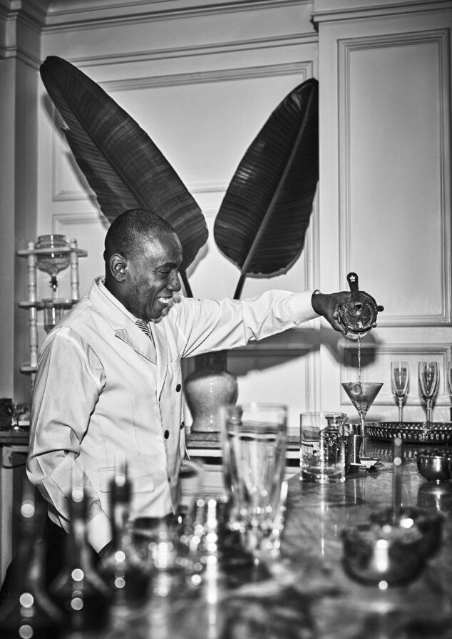 An Ocean Club, A Four Seasons resort hospitality bar staff in a uniform jacket joyfully pours a cocktail into a martini glass at a bar with large decorative leaves in the background.