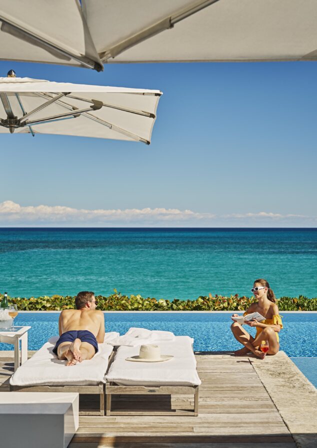 Two people relax near a luxury infinity pool of The Ocean Club, A Four Seasons Resort overlooking the ocean. One is sunbathing on a lounge chair, while the other sits at the pool's edge with a drink under a large hotel umbrella.