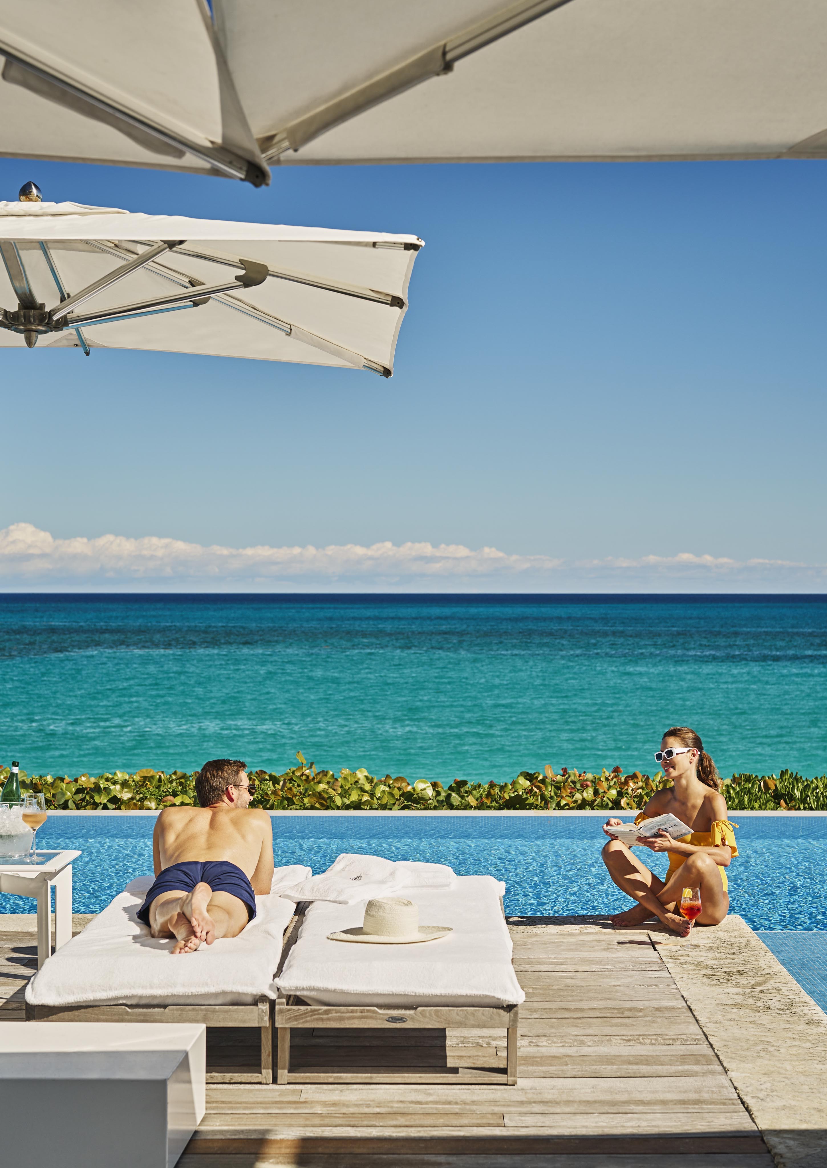 Two people relax near a luxury infinity pool of The Ocean Club, A Four Seasons Resort overlooking the ocean. One is sunbathing on a lounge chair, while the other sits at the pool's edge with a drink under a large hotel umbrella.