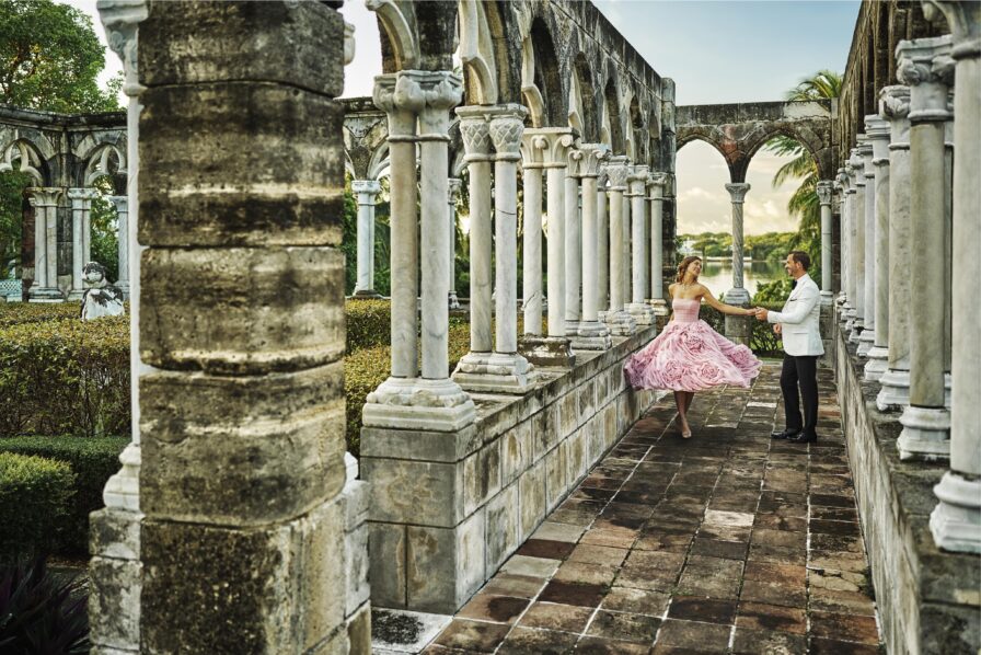 A woman in a pink dress dances with a man in a white suit within a historic French Cloisters of landmark Paradise Island Versailles Gardens found on The Ocean Club, A Four Seasons resort property. The woman wears a pink dress, and the man wears a white jacket. The setting is lush and outdoor.
