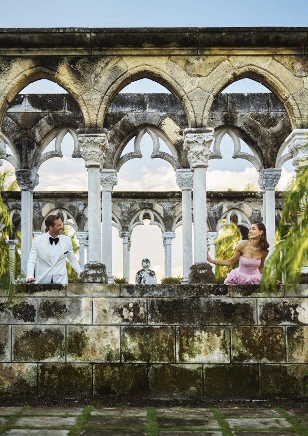 A man in a white suit and a woman in a pink dress stand apart among the historic French Cloisters at the famed Paradise Island Versailles Garden with stone arches and columns, surrounded by greenery.