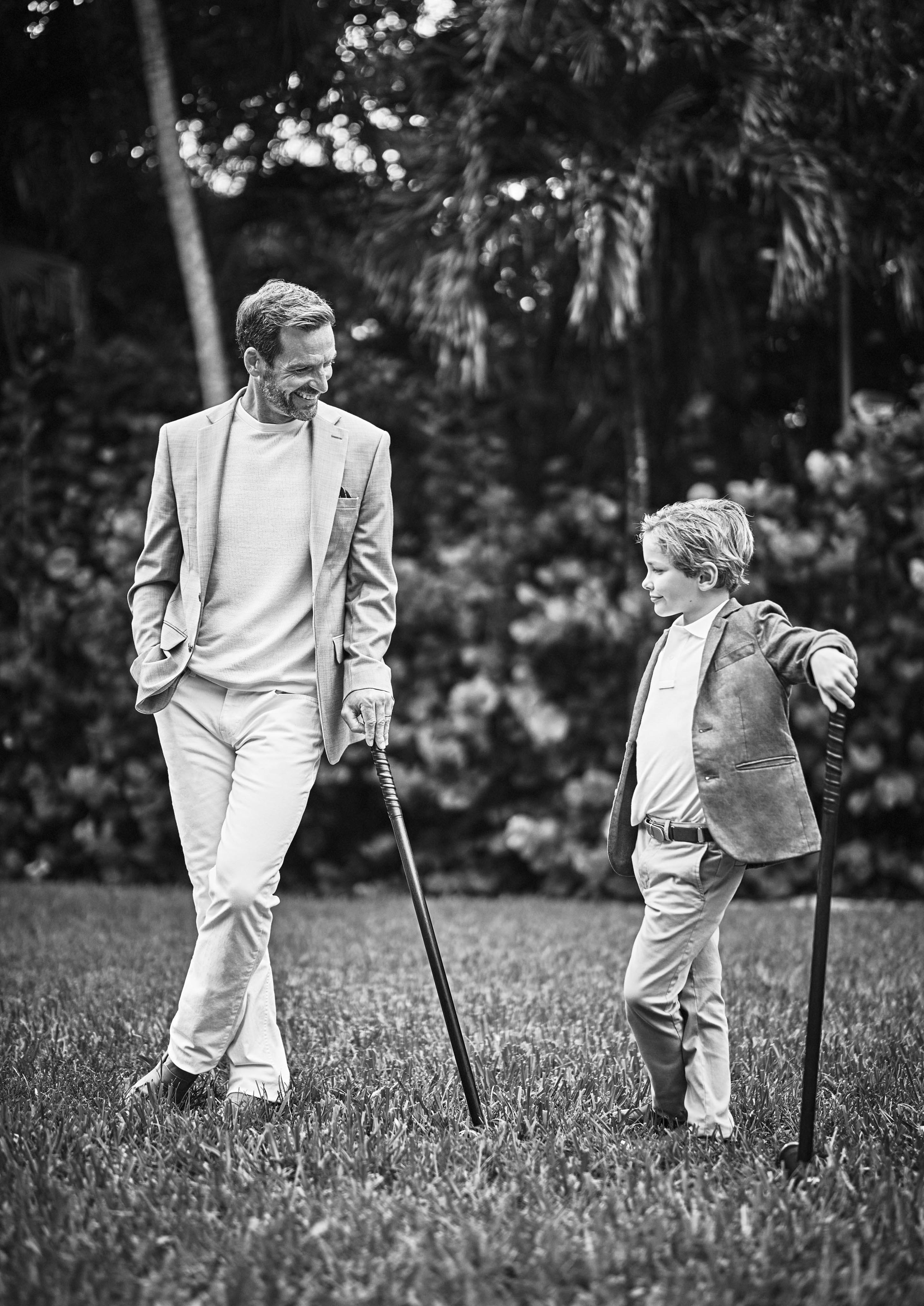 A man and a child in suits walk on landmark Paradise Island Versailles garden , each leaning on their own croquet mallets. black and white image