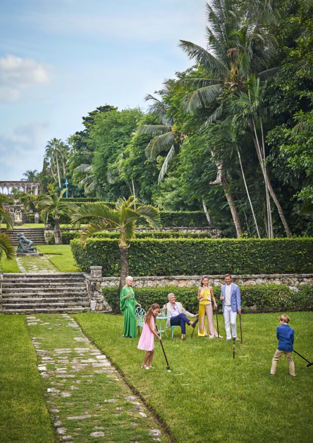 A group of people, dressed in colorful outfits, play croquet on a manicured lawn of the historic Versailles garden within the Ocean Club, A Four Seasons resort luxury property surrounded by tropical trees and stone steps.