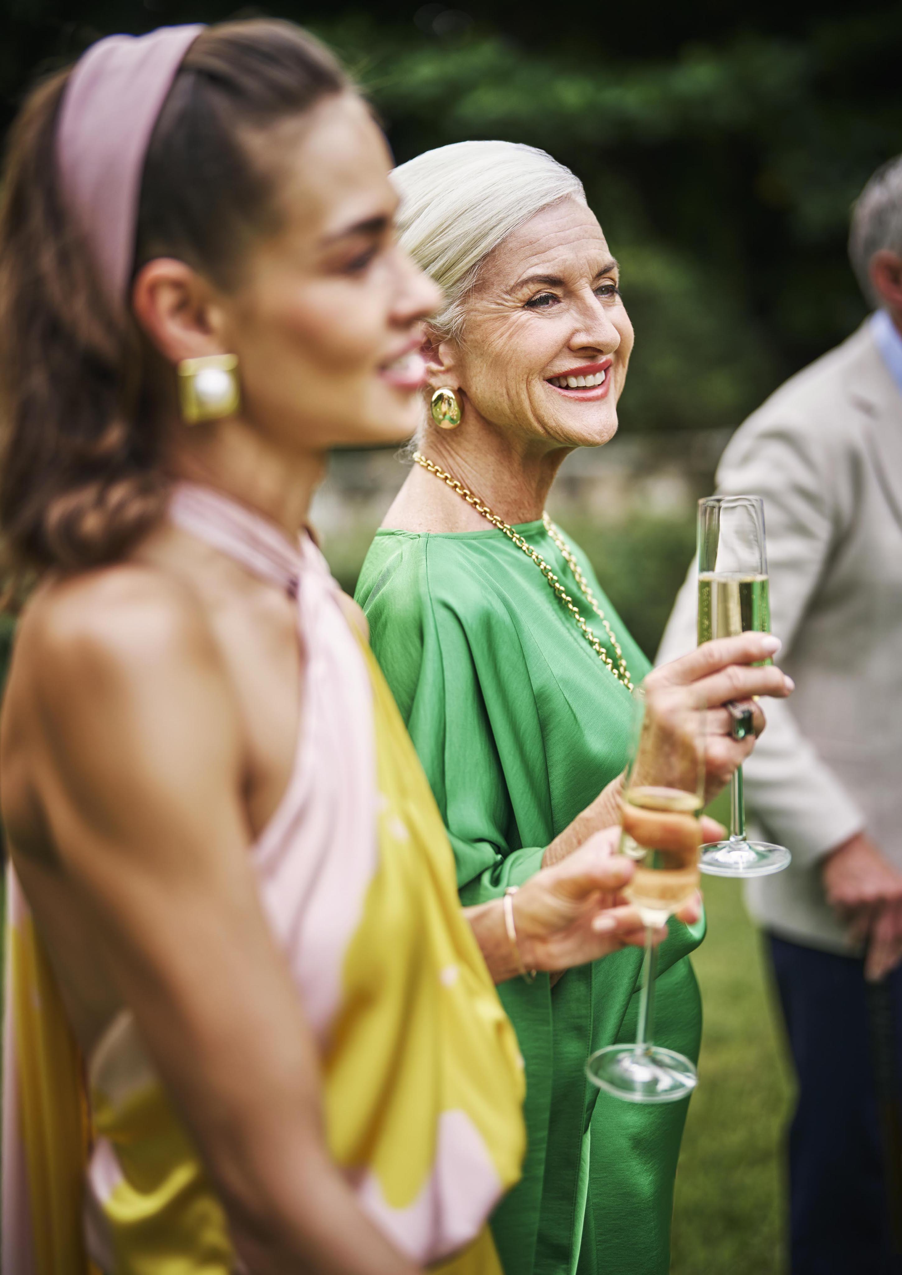 Two women elegantly dressed in bright outfits hold champagne glasses at an outdoor gathering, smiling and engaged on the landmark Ocean Club, A Four Seasons resort Versailles garden property.