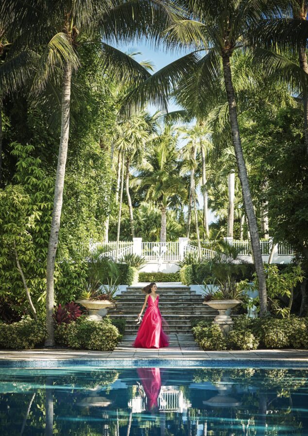 A person in a bright pink dress walks down stone steps leading to the landmark Ocean Club, A Four Seasons resort Versailles pool surrounded by lush greenery and tall palm trees, with a pool and reflection in the foreground.