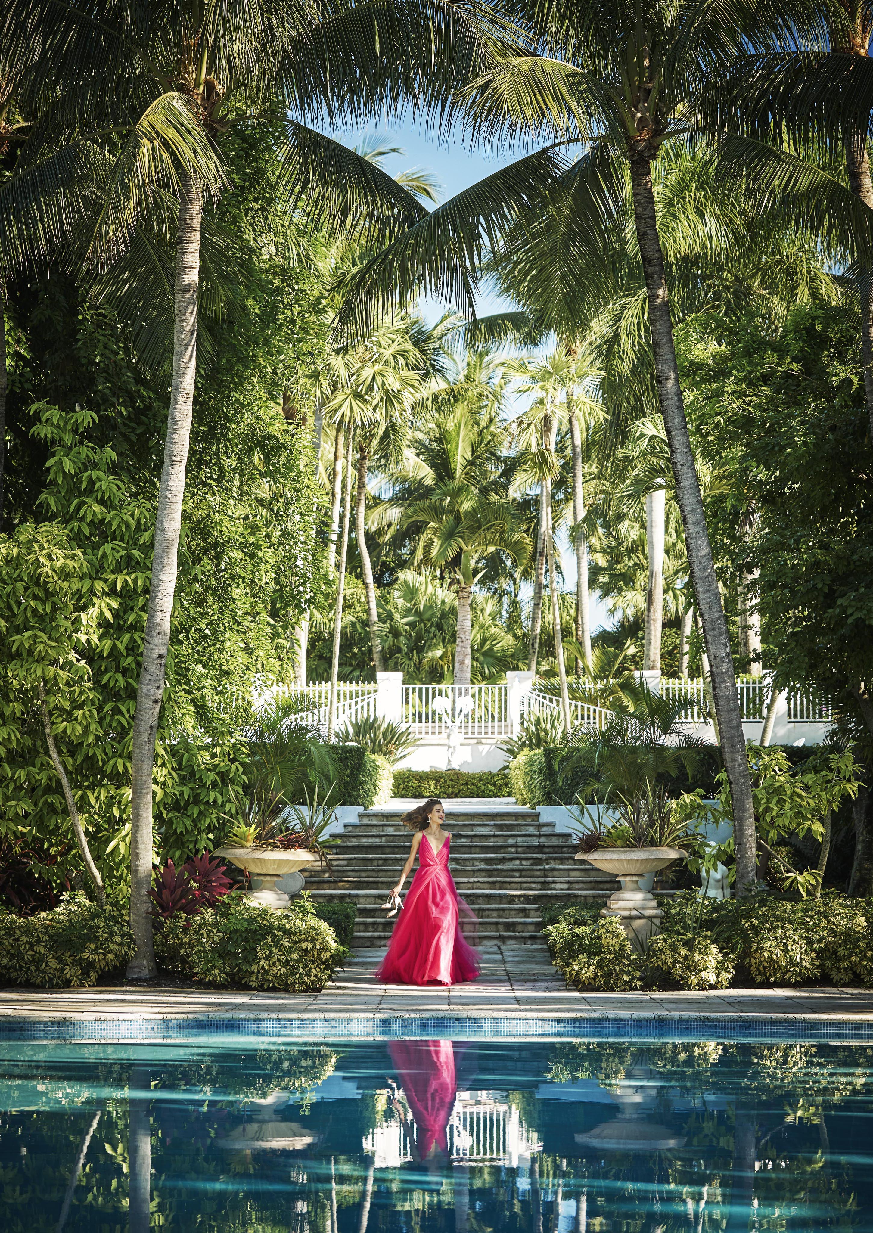 A person in a bright pink dress walks down stone steps leading to the landmark Ocean Club, A Four Seasons resort Versailles pool surrounded by lush greenery and tall palm trees, with a pool and reflection in the foreground.