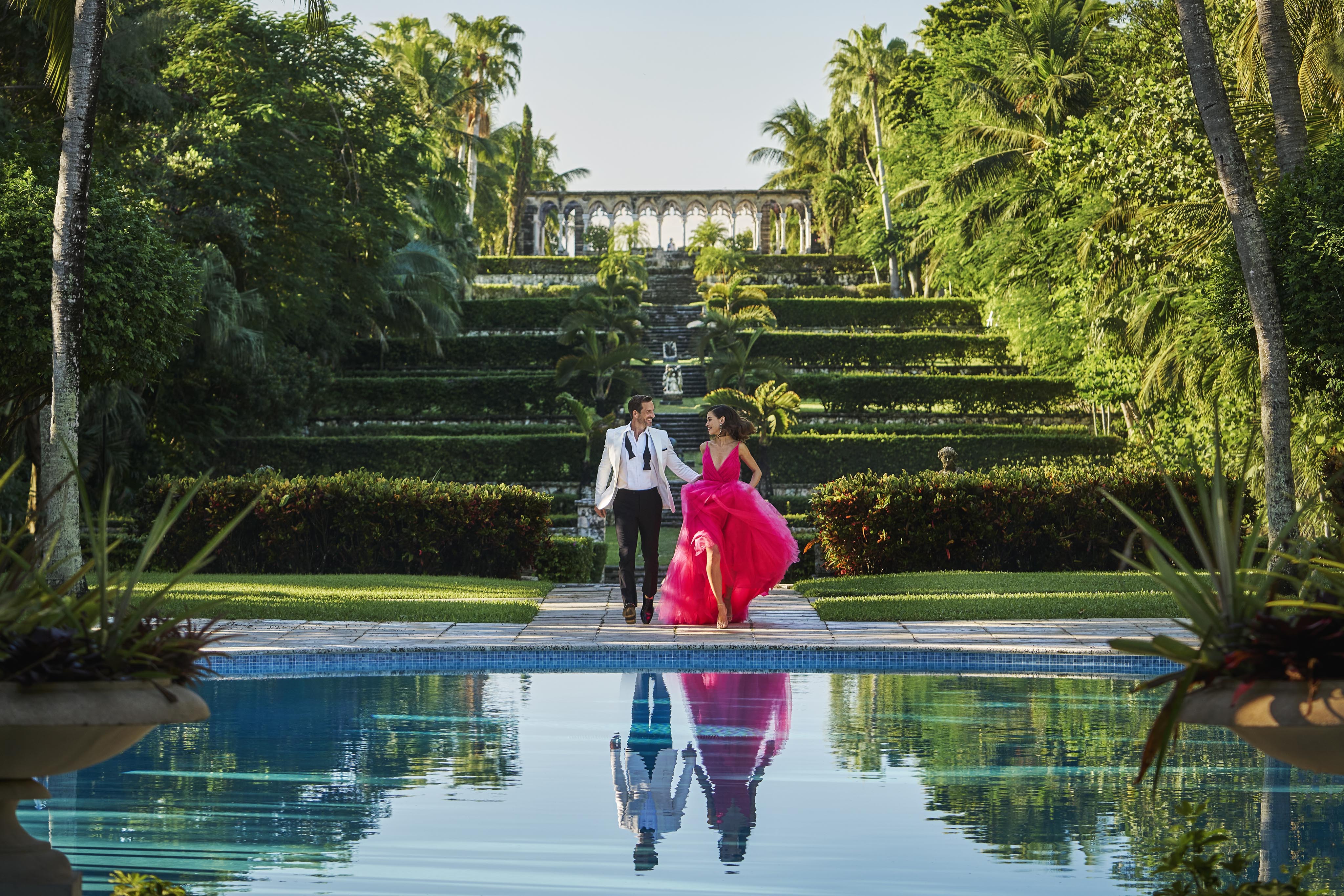 A couple walks hand in hand beside the Ocean Club, A Four Seasons resort pool, surrounded by palm trees and the historic lush Paradise Island Versailles Gardens.The woman wears a bright pink dress, and the man is in a black and white suit.