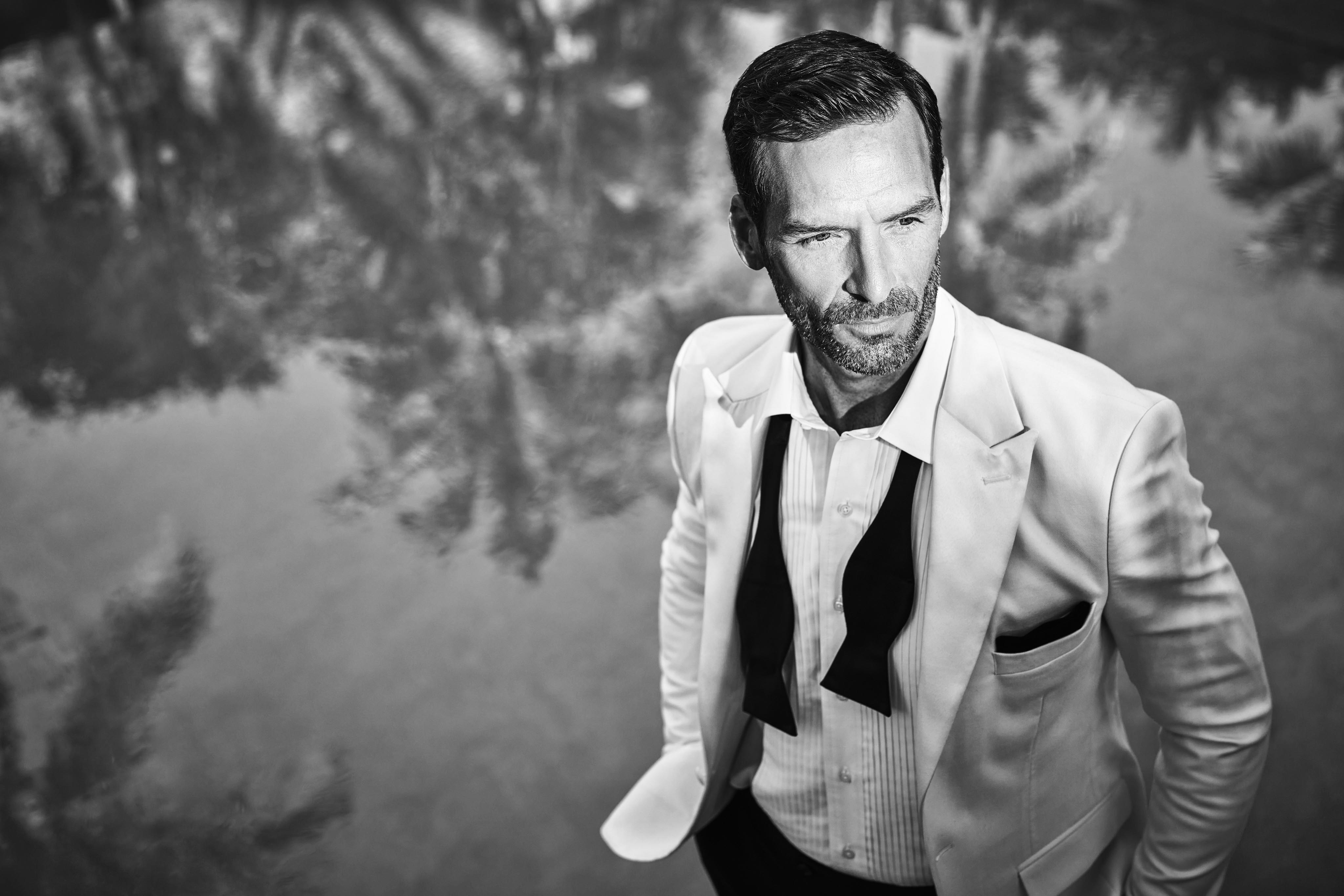 A man in a suit with an untied bow tie stands by the reflective body of water of the historic Ocean Club, A Four Seasons resort Versailles pool, with palm trees visible in the reflection. Black and white image.