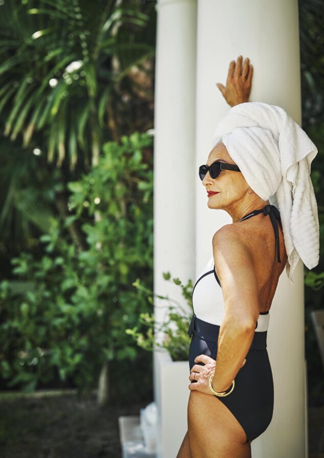 Woman in a luxury brand black and white swimsuit, wearing sunglasses and a towel on their head, stands by a pillar near the Ocean Club, A Four Seasons resort Versailles pool, surrounded by lush greenery.